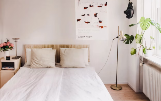 A minimalist bedroom features a double bed with two duvets, crisp white bedding, and two pillows. A bedside table adorned with flowers complements the floor lamp and large poster on the wall, while a lush plant thrives by the window.
