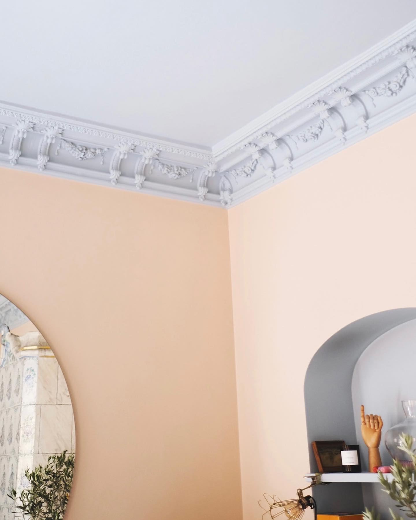 A corner bathed in Toniton Naturally-Pigmented Paint, featuring a peach hue with ornate white crown molding. There’s a circular mirror, an arch niche shelf adorned with various objects like a mannequin hand, and a glimpse of the tiled wall in the background.