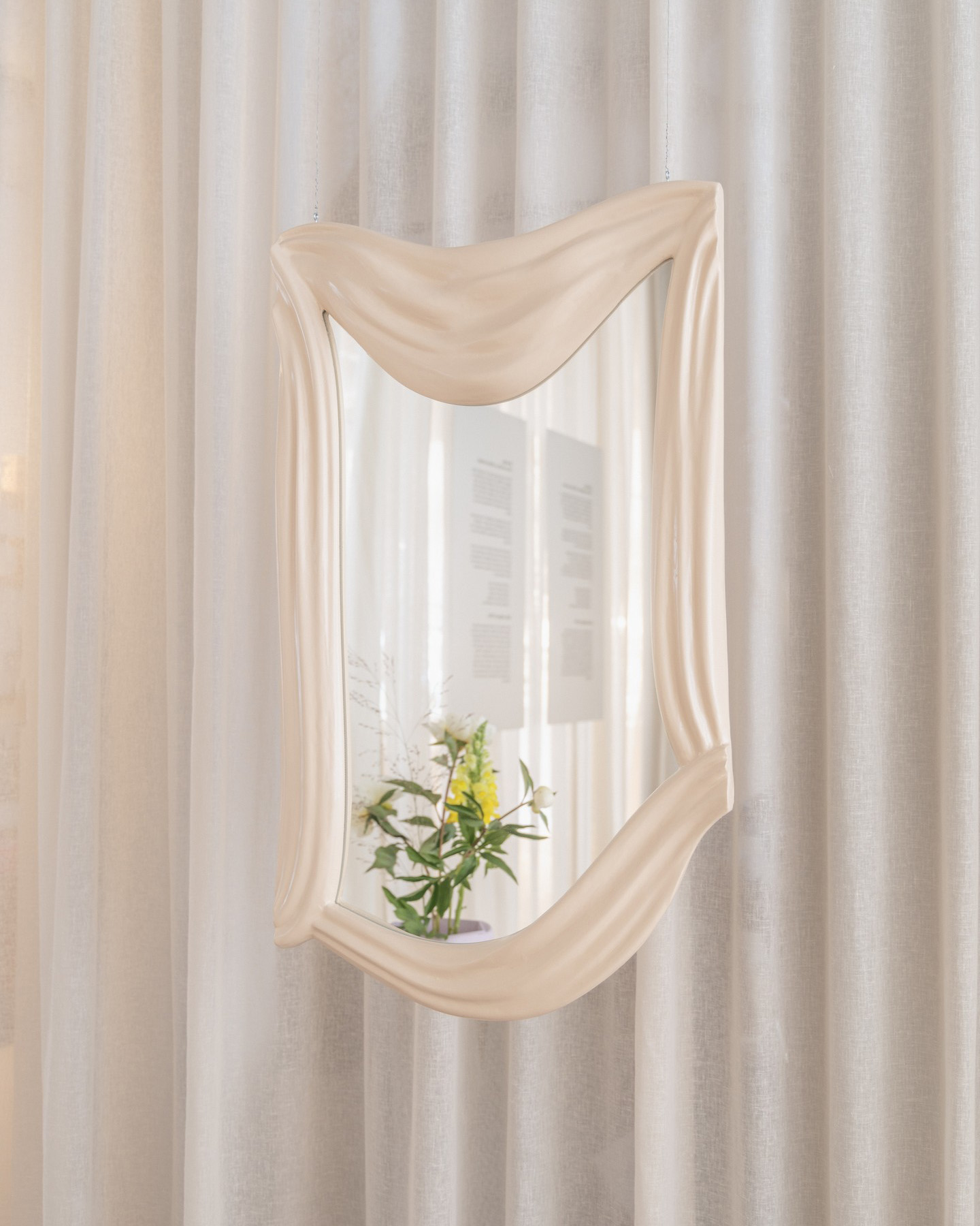 The Alter Ego Mirror, with its large, wavy-edged design and beige frame, perfectly reflects an arrangement of flowers and a lightly textured white curtain. Designed by Ebba Lindgren.