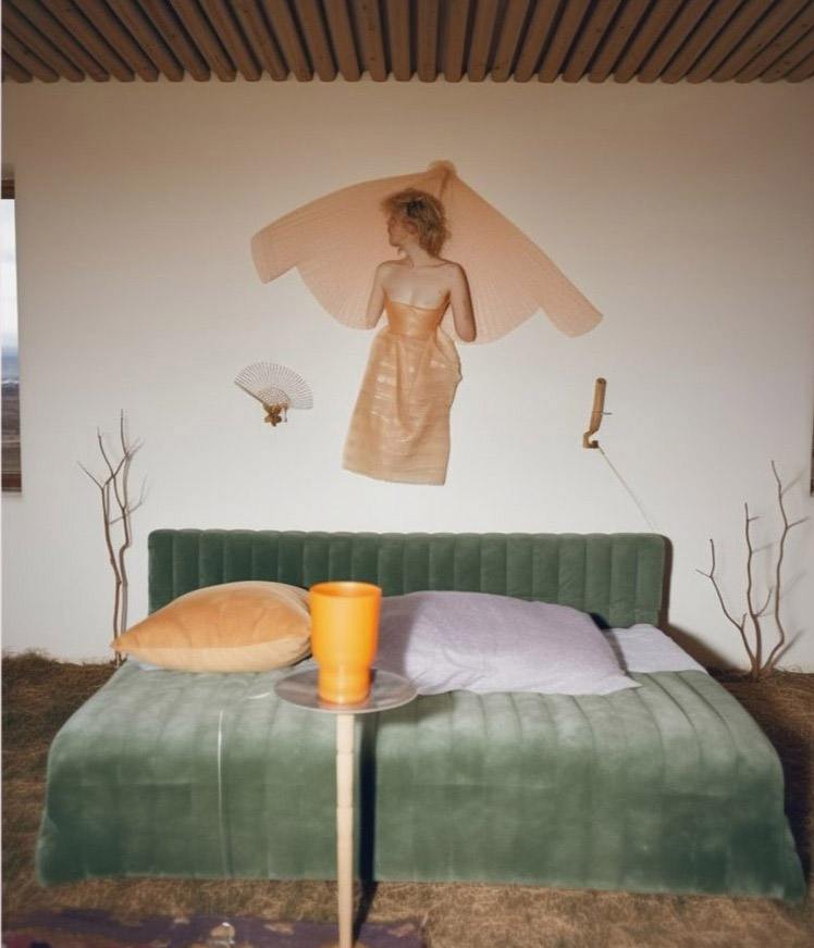 A person in a distinctive gown floats on a wall above a green velvet bed with pillows. A small, round table with an orange cup and a lamp are in the foreground. Sparse decor includes sticks and a fan, capturing an essence reminiscent of Ebba Lindgren's "No Prompt" image generated by her Artificial Intern with AI.