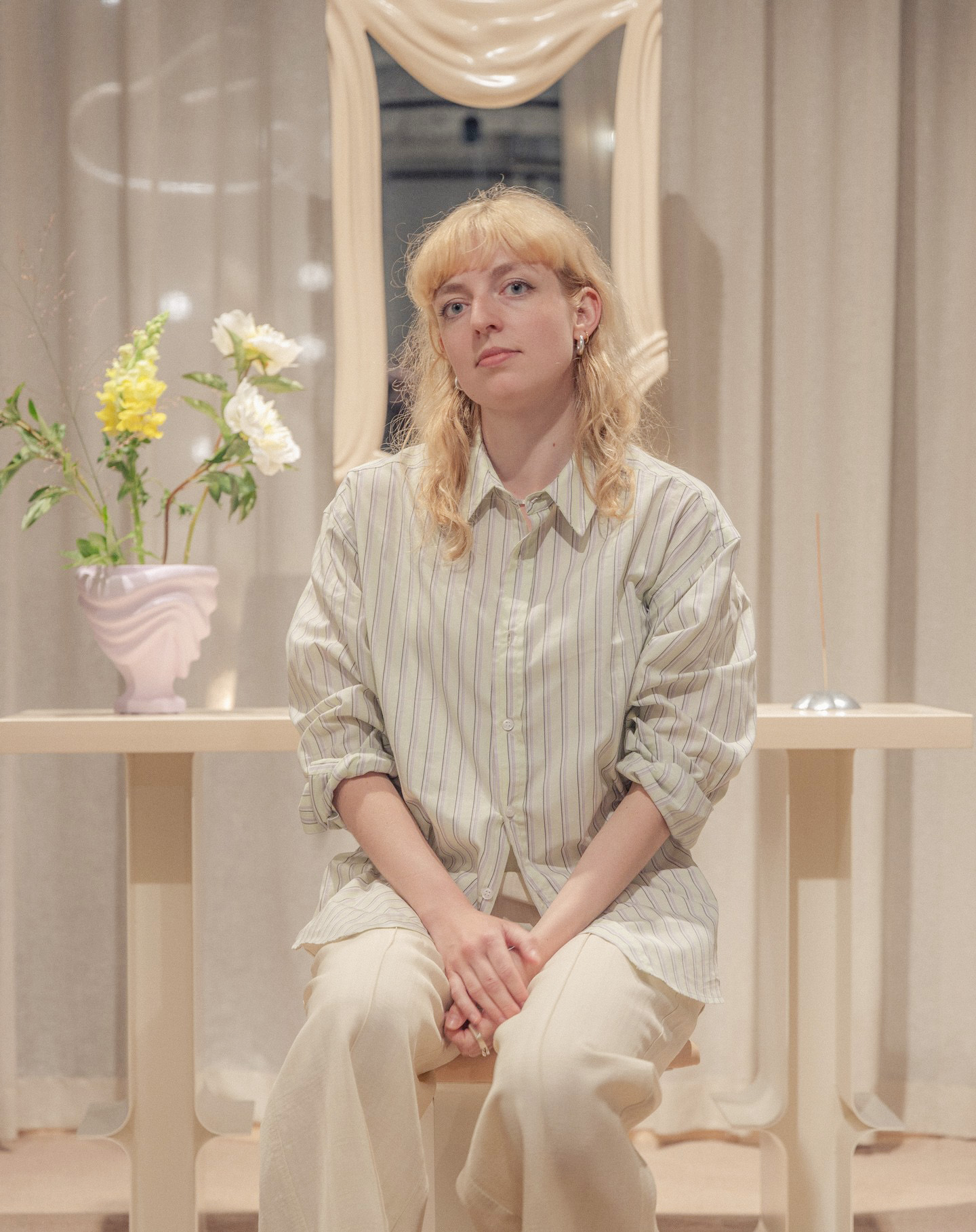 Swedish multidisciplinary artist, Ebba Lindgren, wears a striped shirt and light-colored pants, sits on a chair in front of a table with a vase of flowers and a mirror in the background.