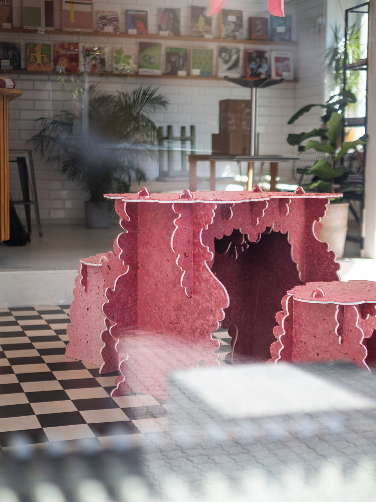 A uniquely designed pink table with matching stools, made from "Really Board" pink textile fiber by Ebba Lindgren, sits in a checkered floor room in the Cowgirl Gallery, Malmo. The background features a bookshelf, potted plants, and a brick wall. The pink textile fibre board is sourced from Kvadrat and Malmö Upcycling Service from textile industry offcuts. This piece is from the "Really Rococo II Exhibition."