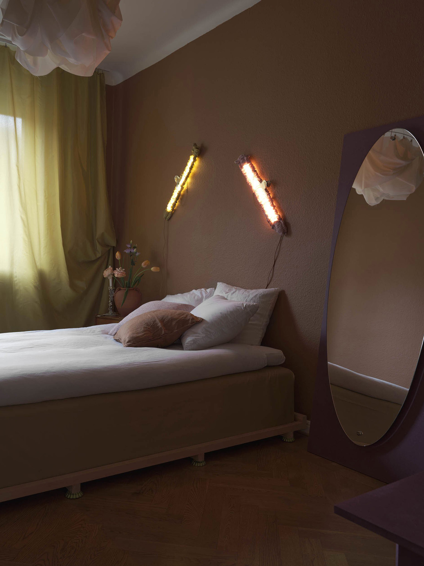 A small bedroom with a brown wall, an oval mirror, a bed with white bedding and brown pillows. Two vertical lights are on the wall. Green curtains partly cover a window. Sparse decorative items evoke the curated simplicity of the "Scenes From a Home" exhibition by Ebba Lindgren.