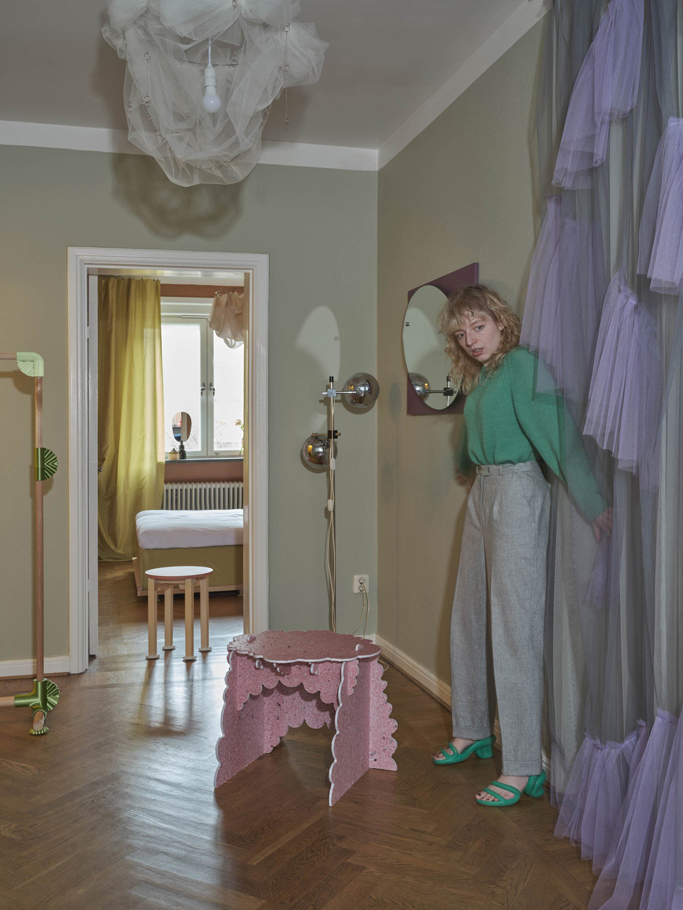 A woman stands in a stylish room with green walls, wooden flooring, a pink decorative table, and purple tulle curtains. A bedroom with a bed and yellow drapes is visible through an open doorway, reminiscent of an artful gallery display.