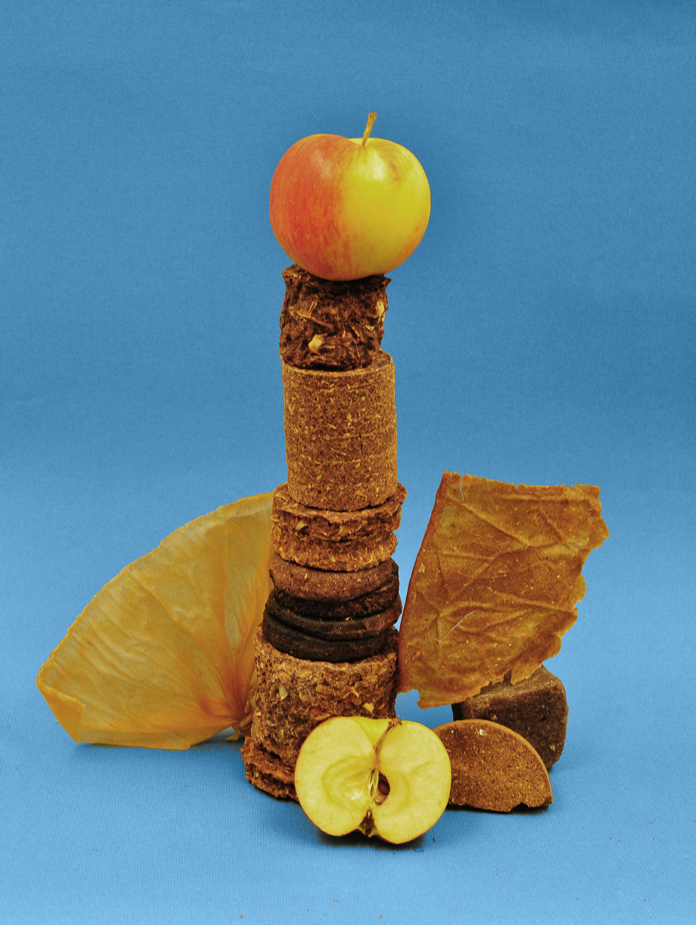 A stack of varied circular objects topped with a whole apple, along with a sliced apple and two large, irregular brown pieces made from crushed apple residue from Kivik's cider press, all set against a blue background.