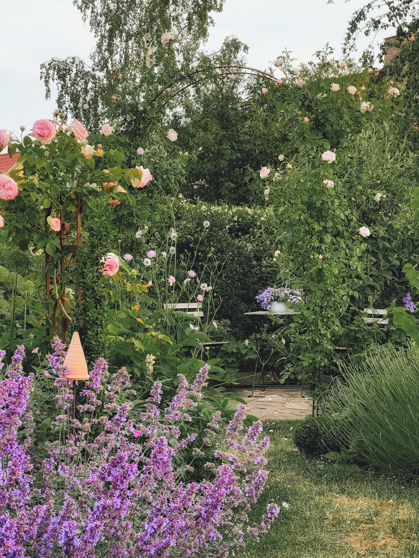 A garden scene with various blooming flowers, including pink roses and purple blossoms, surrounding a small patio with a table and chairs, all under an archway of greenery.