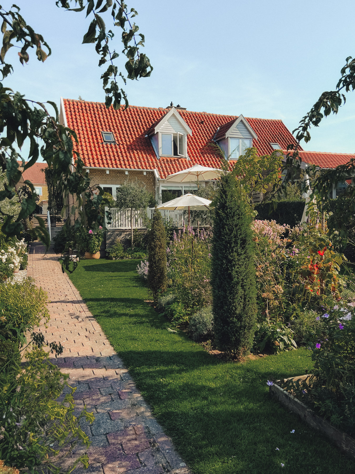 A well-maintained garden with a brick path leads to a house with a red-tiled roof. The garden features trees, bushes, and colorful flowers, with an outdoor seating area under an umbrella.