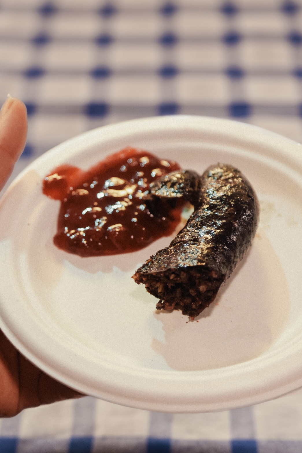A white paper plate holding a partially eaten sausage and a serving of red sauce is seen against a blue and white checkered tablecloth background.