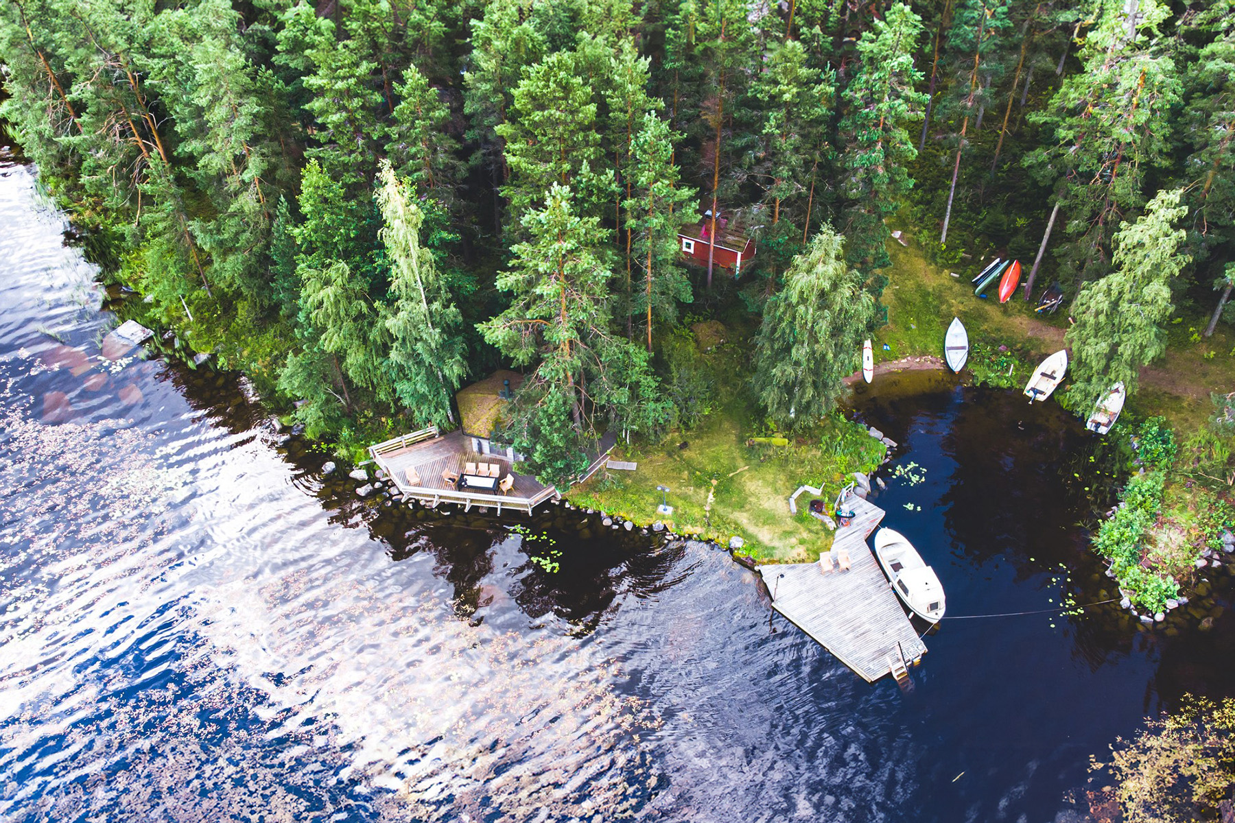 Aerial view of a forested lakeshore with boats docked at a small pier, a luxurious red cabin surrounded by dense trees, and outdoor seating areas. This Taivasalla gem offers an open sky experience, embodying the best of autumn in Finland. Perfect for those seeking the best hotels near Tampere.