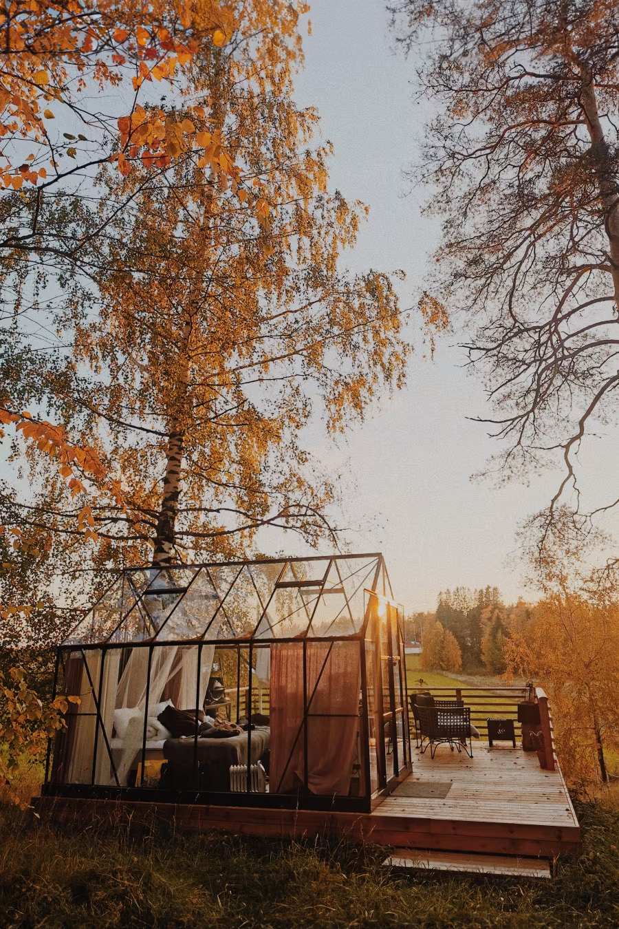 A glass greenhouse, known as the Taivasalla Glamping Glass Cabin, surrounded by autumn trees stands on a wooden deck at sunset. This luxurious retreat includes seating arrangements and overlooks a grassy field by the lakefront. Experience one of Tampere's best hotels under an open sky in Autumn Finland.