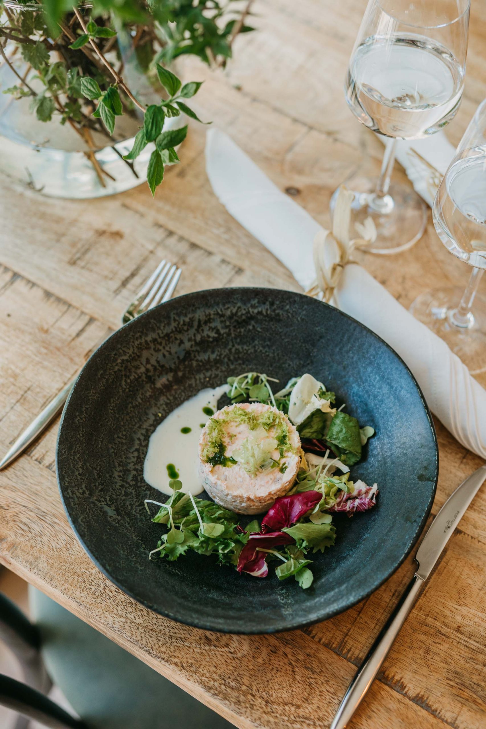 A black plate with a gourmet dish featuring diced fish or seafood on a creamy sauce, garnished with greens and microgreens, placed on a wooden table next to cutlery, glasses, and a white napkin.