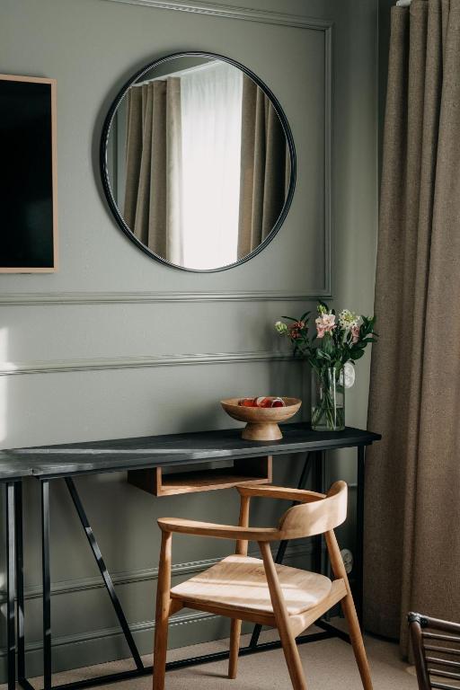 A wooden chair is placed in front of a black desk against a wall with a round mirror and a rectangular frame. A vase with flowers and a bowl with fruit are on the desk. Curtains cover the window.