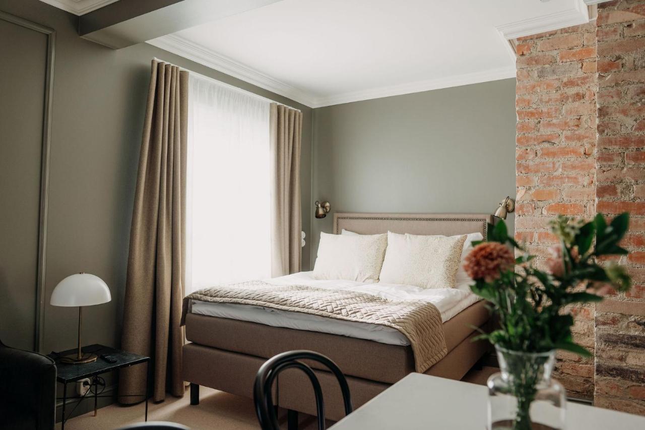 A bedroom with a double bed, beige bedspread, and light-colored curtains. There is a brick accent wall, a bedside table with a lamp, and a vase with flowers on a table in the foreground.