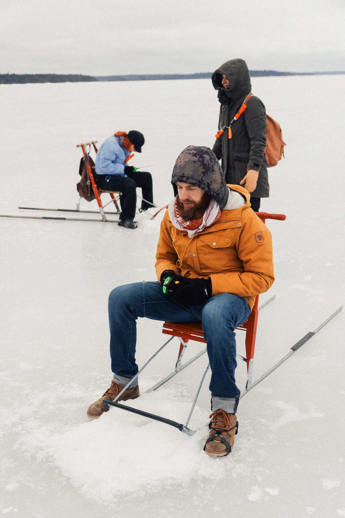 Three people ice fishing on Frozen Lake Näsijärvi near Siilinkari Light House. The foreground shows a person in an orange jacket sitting on a chair, focusing on fishing. Two others are in the background, also engaged in ice fishing.