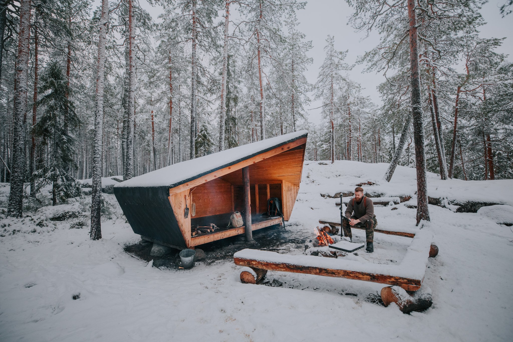 A person sits by a campfire near a small, modern wooden lean-to shelter in the snowy, serene forests of Kintulammi Nature Reserve, surrounded by tall pine trees.