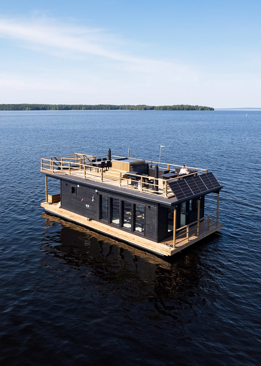A modern houseboat with a wooden deck is floating on a calm lake under a clear blue sky in Tampere. The houseboat features large windows, a rooftop sitting area, and even includes party sauna for an unforgettable experience.