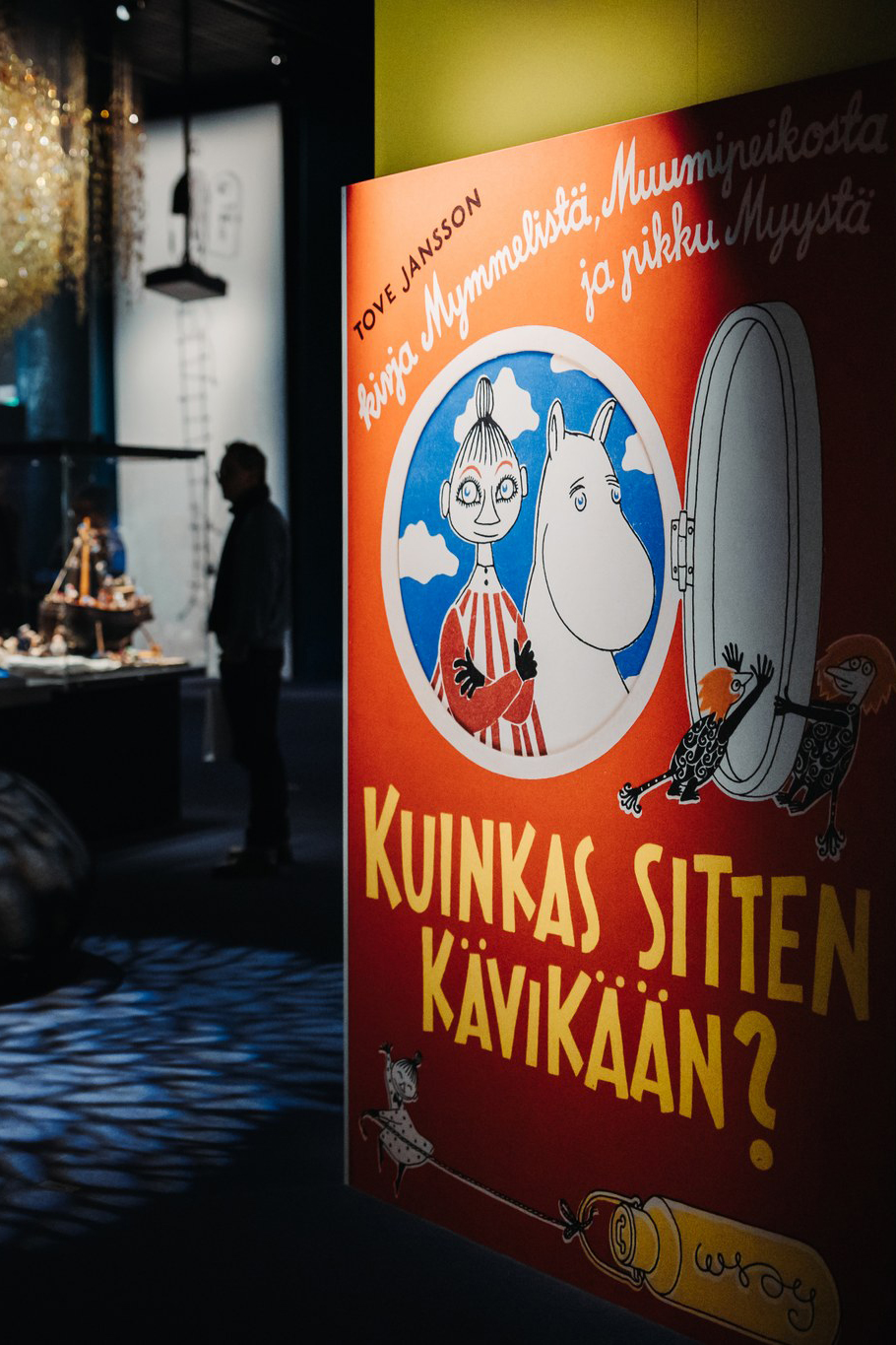 A poster with Moomin characters and Finnish text is displayed in a dimly lit exhibition space at the Moomin Museum in Tampere, Finland, with a person and various exhibits visible in the background.