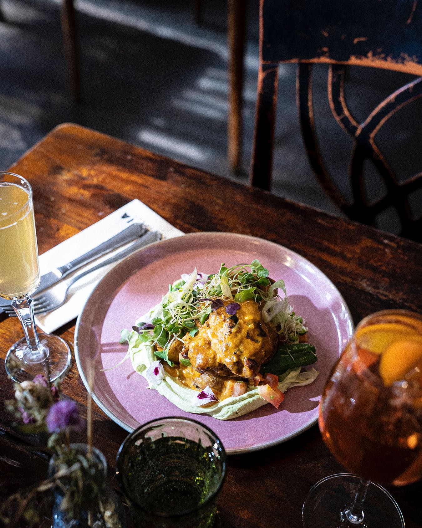 A plate with a dish of vegetables and sauce, accompanied by two glasses of beverages, is placed on a wooden table with utensils.