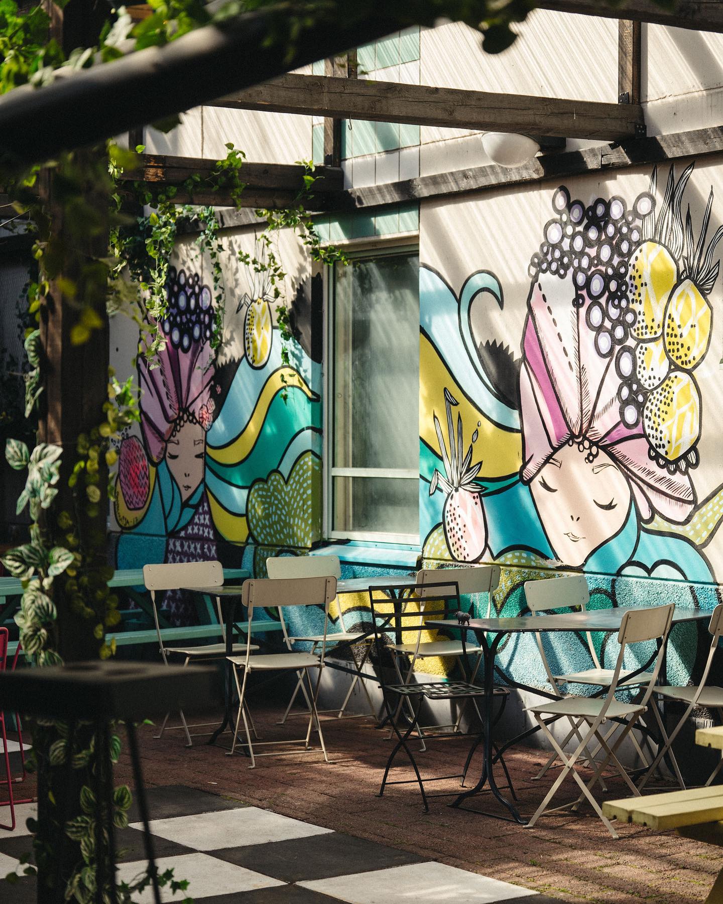 Outdoor seating area with metal chairs and tables, featuring colorful wall murals depicting abstract faces and fruit motifs. The area is decorated with hanging plants, bathed in sunlight, offering a charming ambiance reminiscent of Tampere, Finland.