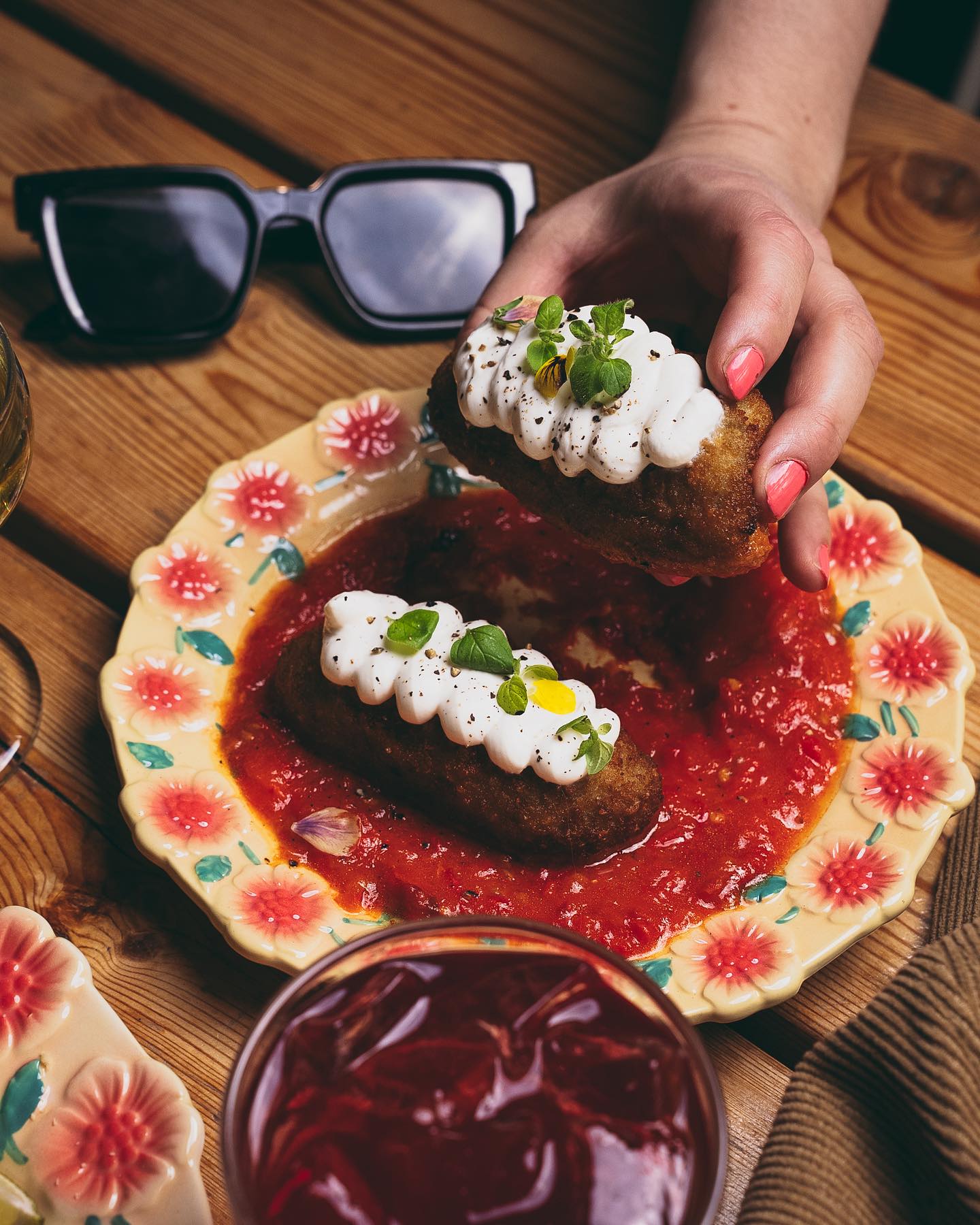 A person holds a croquette topped with cream and herbs above a plate filled with tomato sauce, with sunglasses on the table.