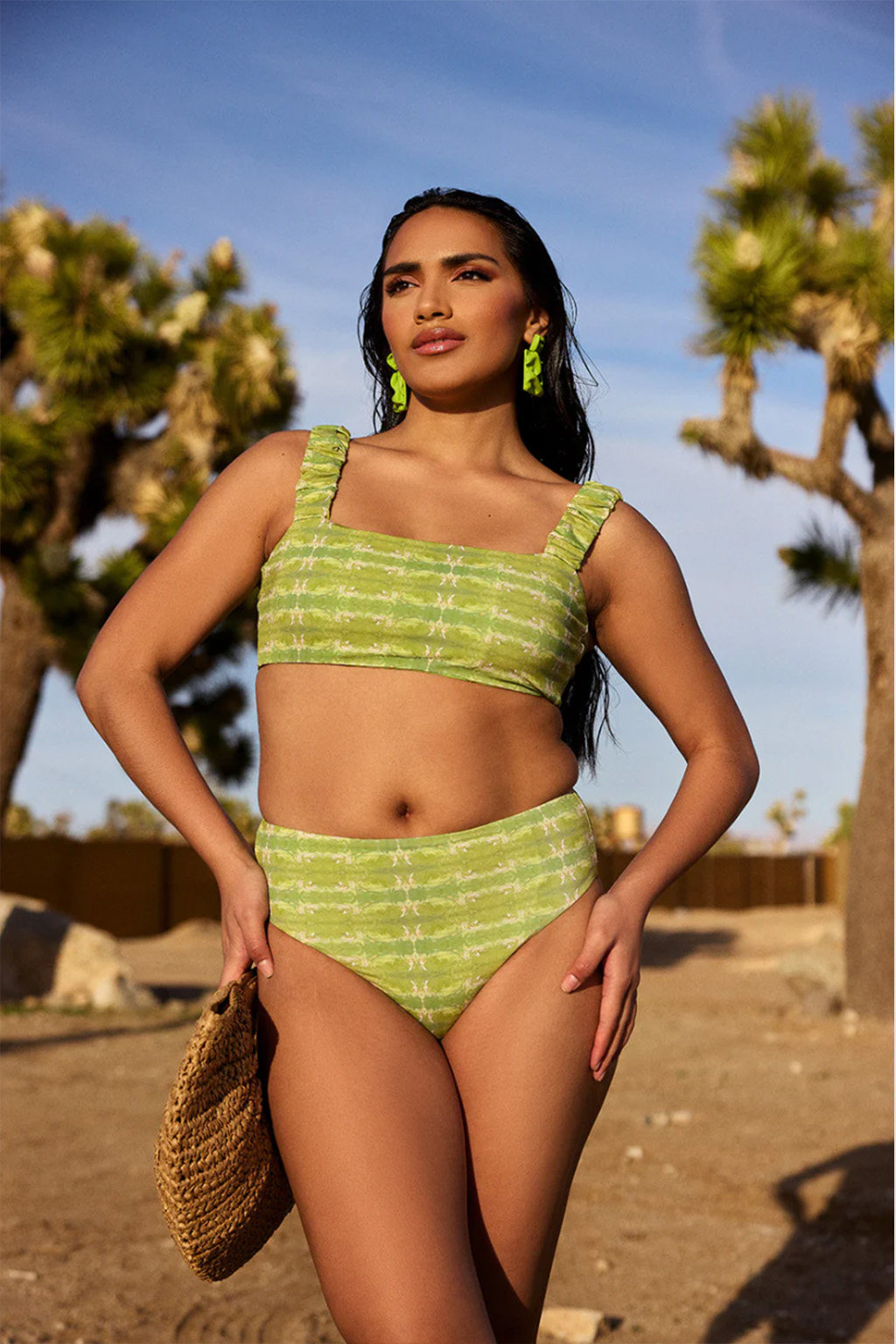 A person stands outdoors wearing a stunning lime green bikini from Sauna Nomads Swimwear, holding a woven bag, with trees and rocks in the background. This scene perfectly captures the essence of the best swimsuits for summer.