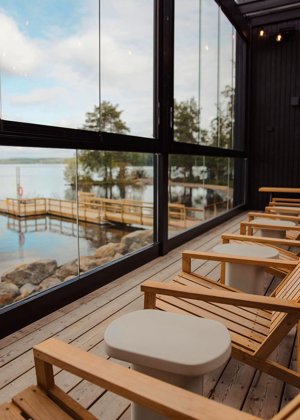 A sauna in a wooden lounge chairs and tables on a covered outdoor deck with large glass panels, overlooking a lake with a dock and trees in the background.