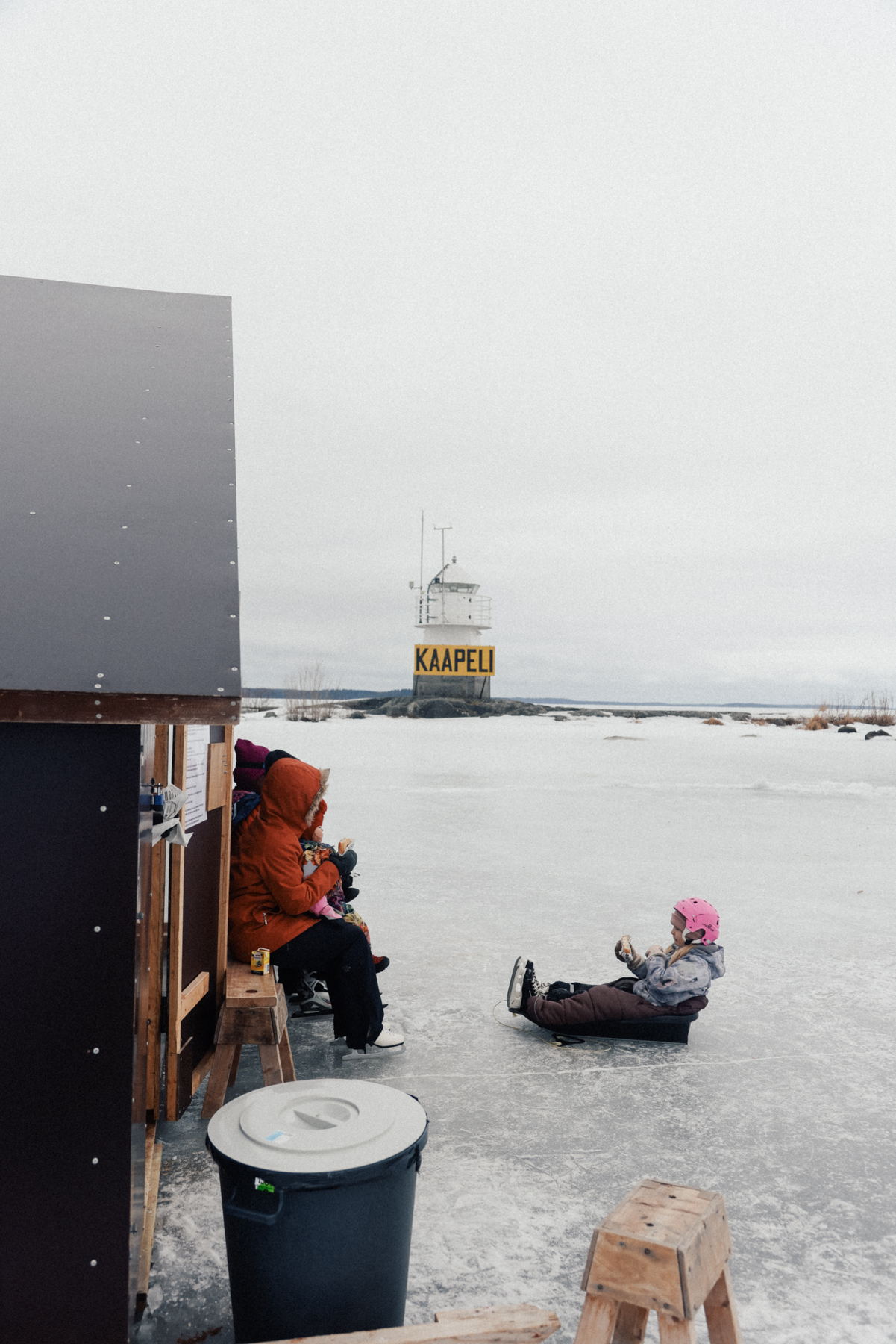 People enjoying food from a cafe on the ice sit outside a small structure on frozen Lake Näsijärvi, near the Siilinkari Lighthouse with the word "KAAPELI" in the background. One child is sitting in a sled on the ice next to a black barrel.