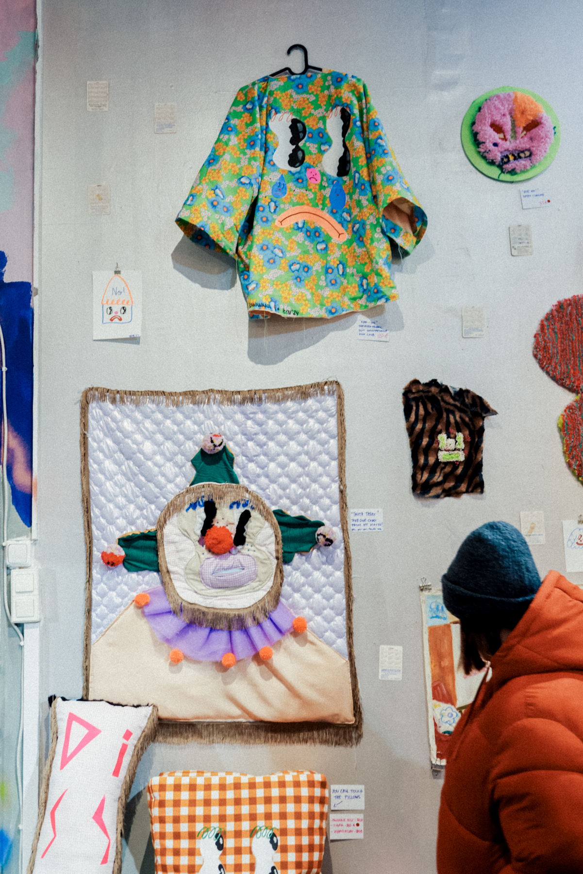 A person in an orange jacket observes a wall displaying colorful, quirky textile art, including a hanging floral-patterned Pispala Clothing jacket, framed fabric pieces, and various textile crafts. This eclectic display showcases the vibrant artistic clothing scene in Tampere.