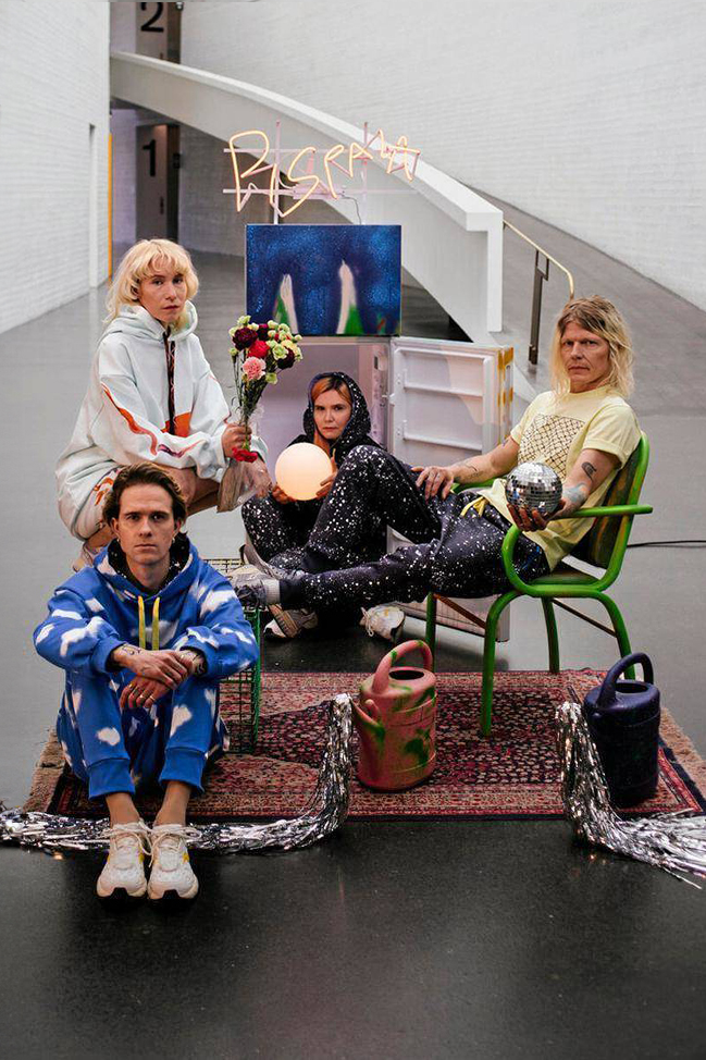 Four people in colorful, eclectic outfits pose in an art gallery setting with various objects, including watering cans, a lamp, and flowers. A neon sign and framed artwork are in the background, showcasing the artistic vibe of Pispala Clothing from Tampere.