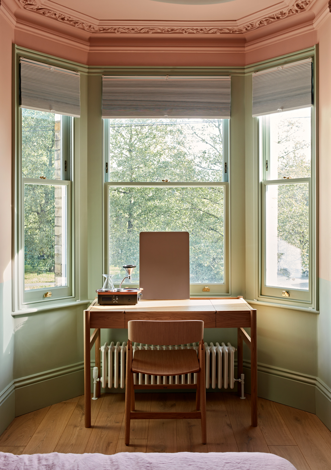 A wooden desk with a chair located in front of three tall windows in a room painted with Toniton Naturally-Pigmented Paint, featuring green and pink hues. The windows offer a view of an outdoor scene with trees.