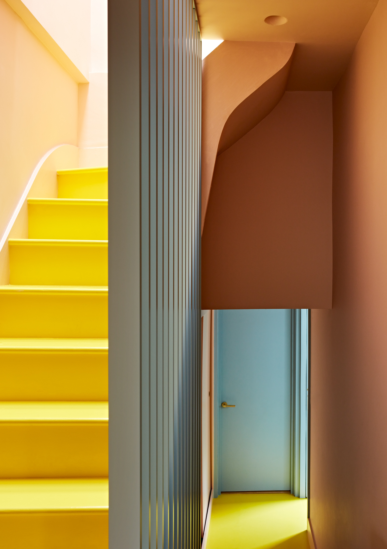 A narrow hallway with pastel-colored walls, a blue door, and bright yellow stairs features natural paint. The stairs and hallway have a modern, minimalist design with clean lines and soft light from above.