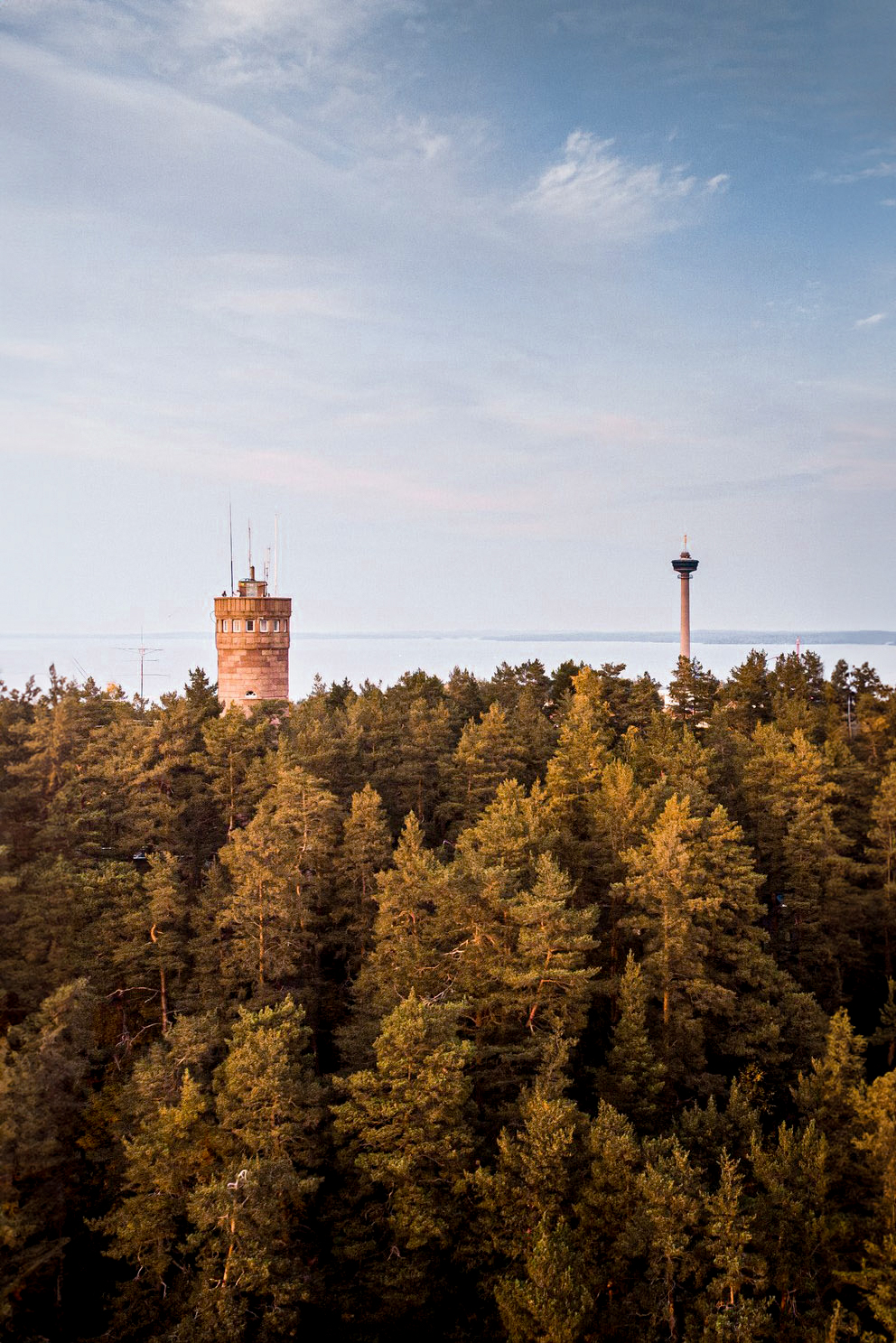 Two towers stand amidst a dense forest under a partially cloudy sky. One tower is the medieval stone Pyynikki Coffee Shop & Observation Tower, while the other is a modern communication tower. Nasinneula can be seen from the ridge along the Pyynikki Nature Reserve Hiking Trail.