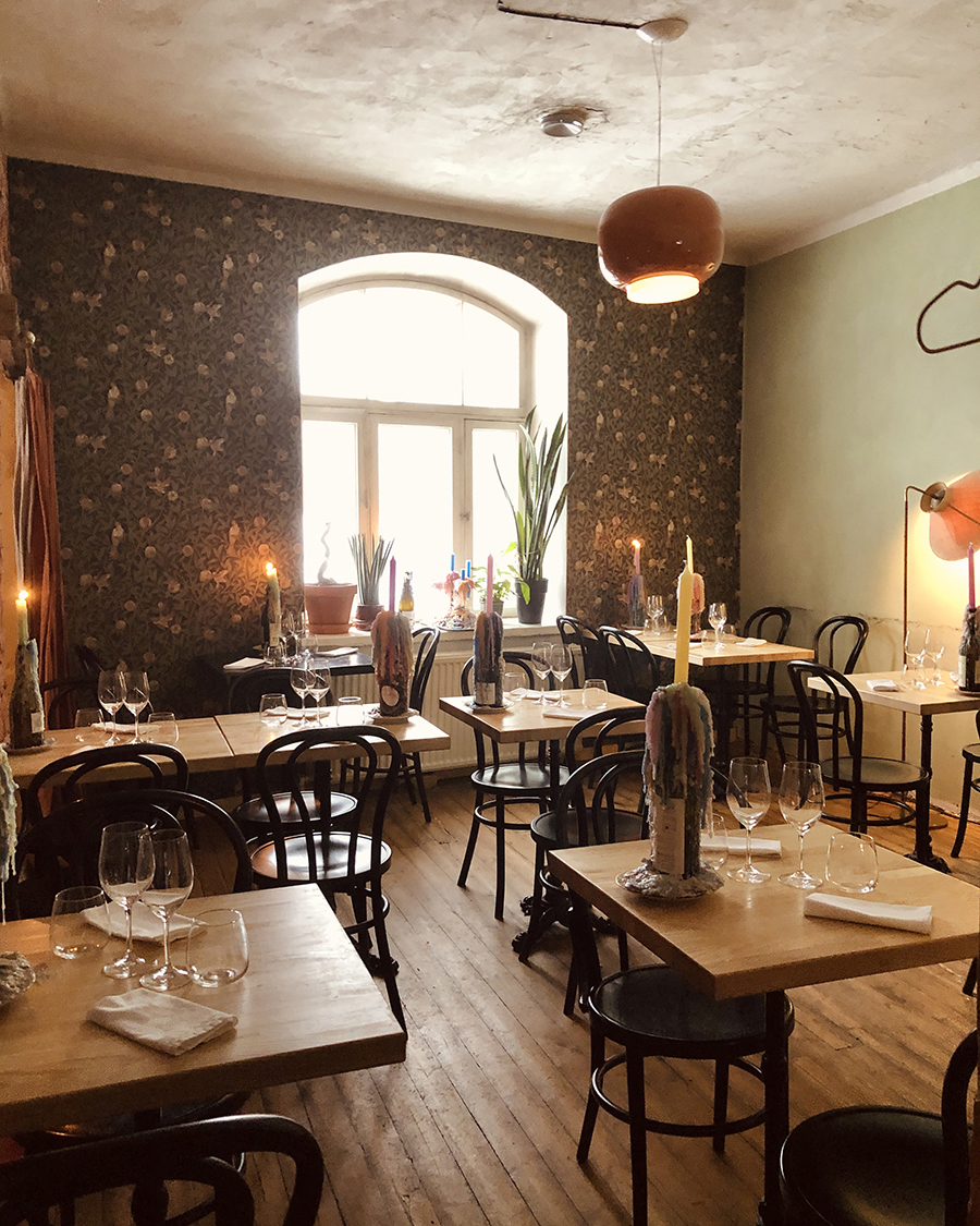 A cozy restaurant interior with wooden tables and chairs, candlelit ambiance, and a large arched window letting in natural light.