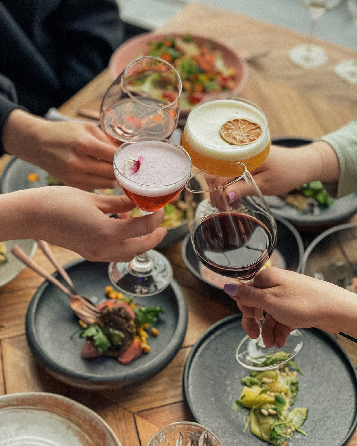 Four hands clink cocktail and wine glasses over a wooden table set with plates of food.