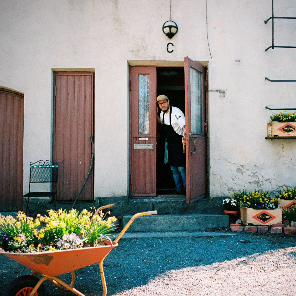 Person standing in a doorway next to a wall with small flower boxes. There is a wheelbarrow filled with flowers in the foreground.