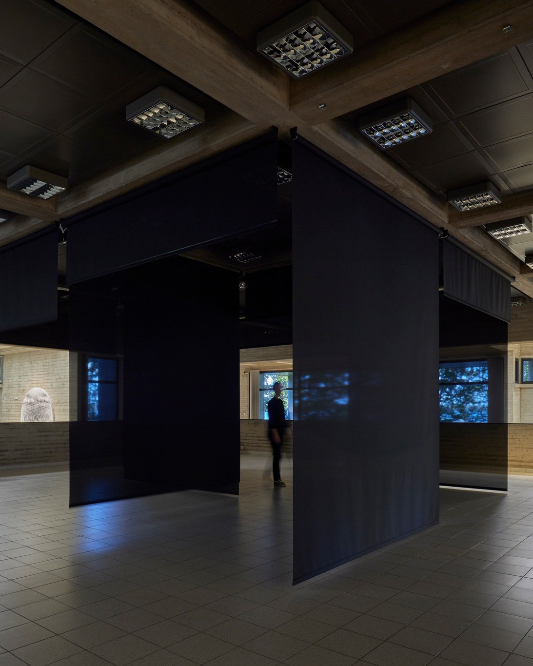 A person is standing in a dimly lit room with black partition panels and tiled flooring inside the Sara Hilden Museum Tampere, surrounded by soft light coming from adjacent spaces.