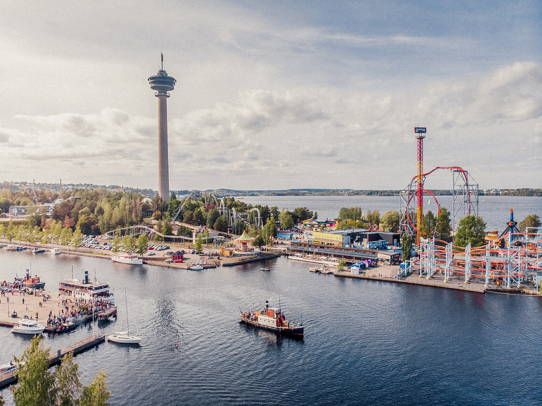 Aerial view of Särkänniemi Amusement Park by a lake, featuring a tall observation tower, colorful rides, boats on the water from the Steamboat Regatta Lakeland Festival, and a mix of trees and buildings. The sky is partly cloudy.