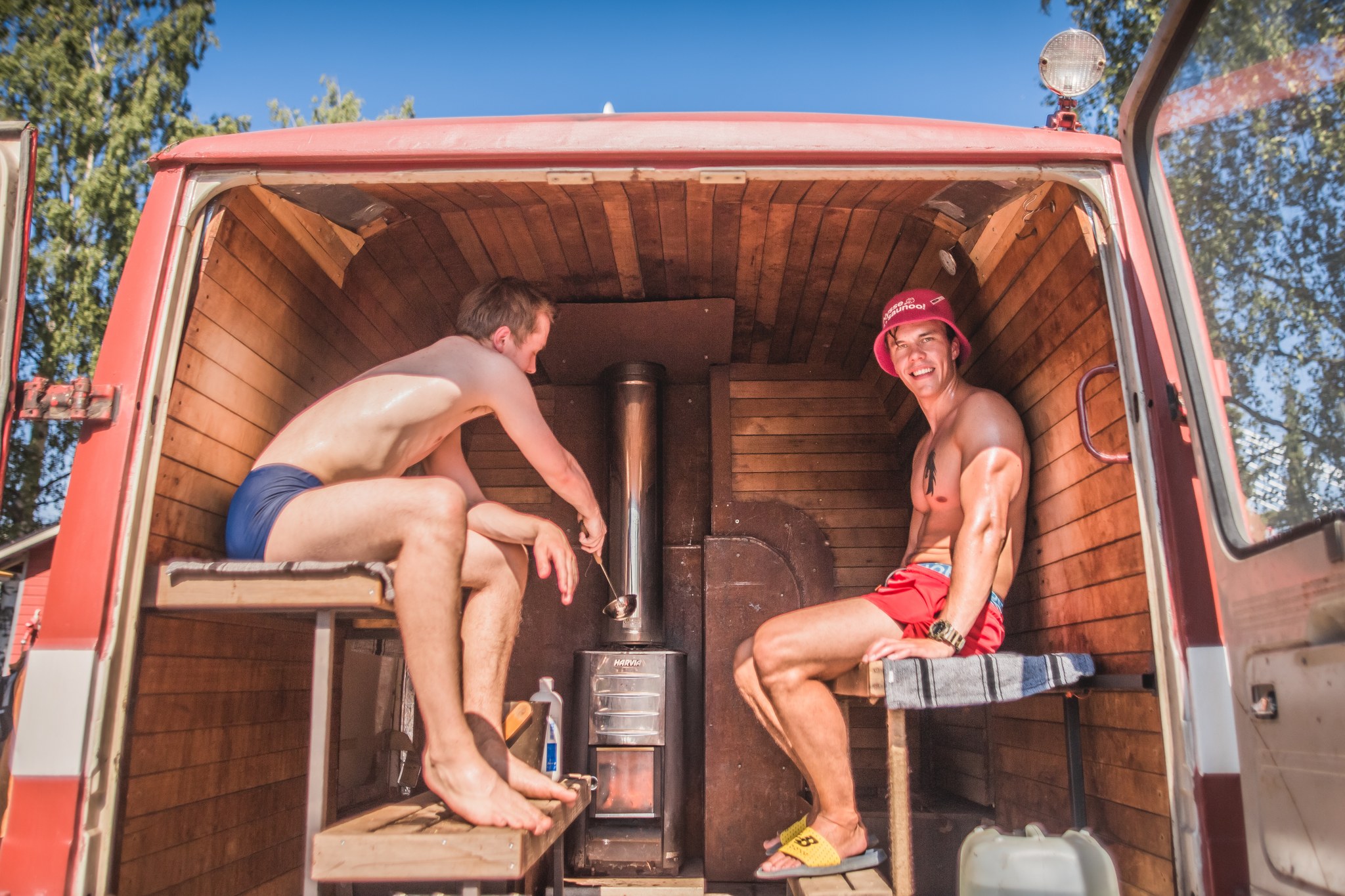 Two men in swimwear sit inside a converted van sauna with a wood-paneled interior, a metal wood stove between them, and a blue sky visible outside.
