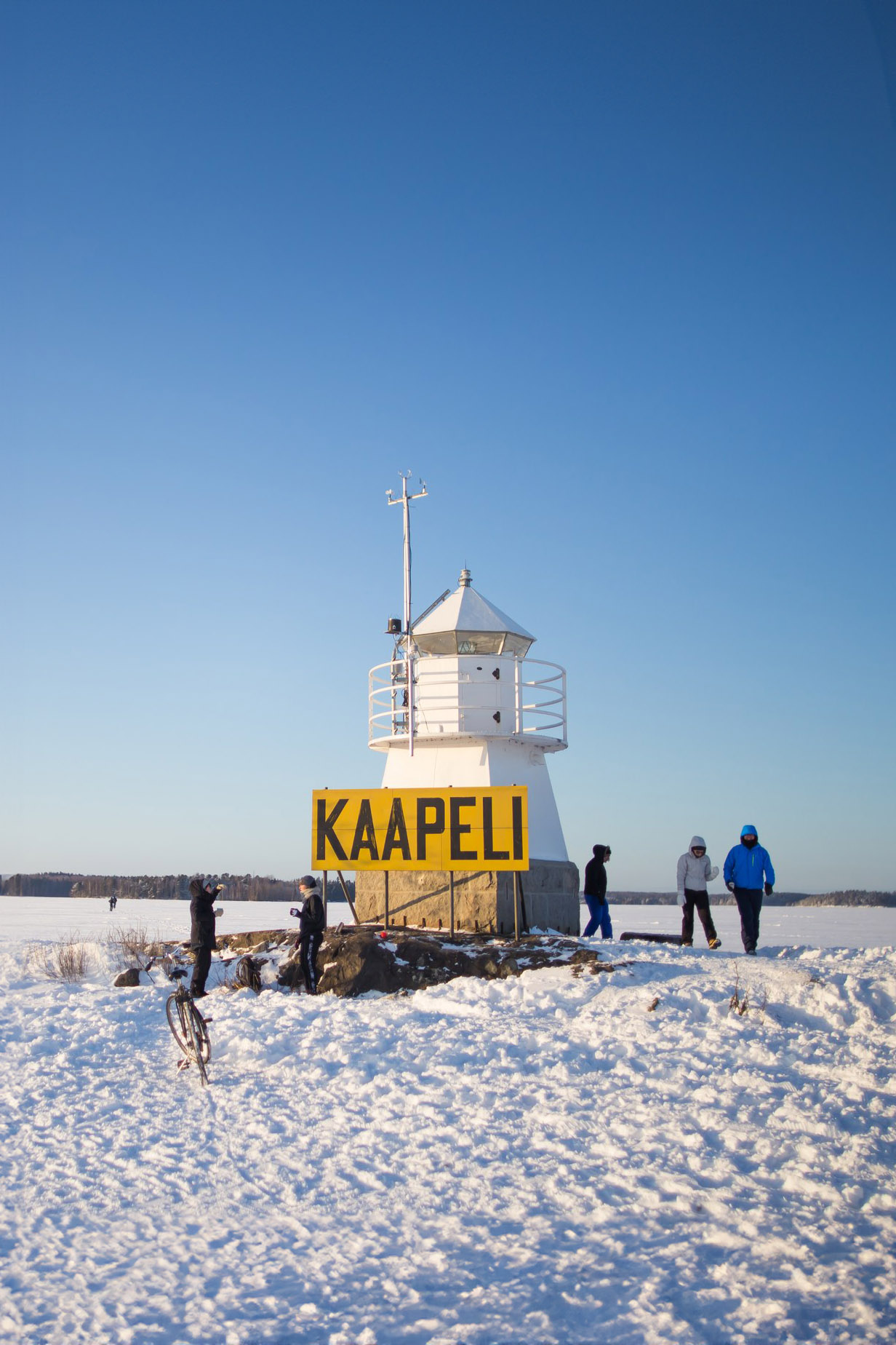 A group of people stand around the Siilinkari Lighthouse in the middle of frozen Lake Näsijärvi, Tampere, with "KAAPELI" written on a sign, set against a snowy landscape.
