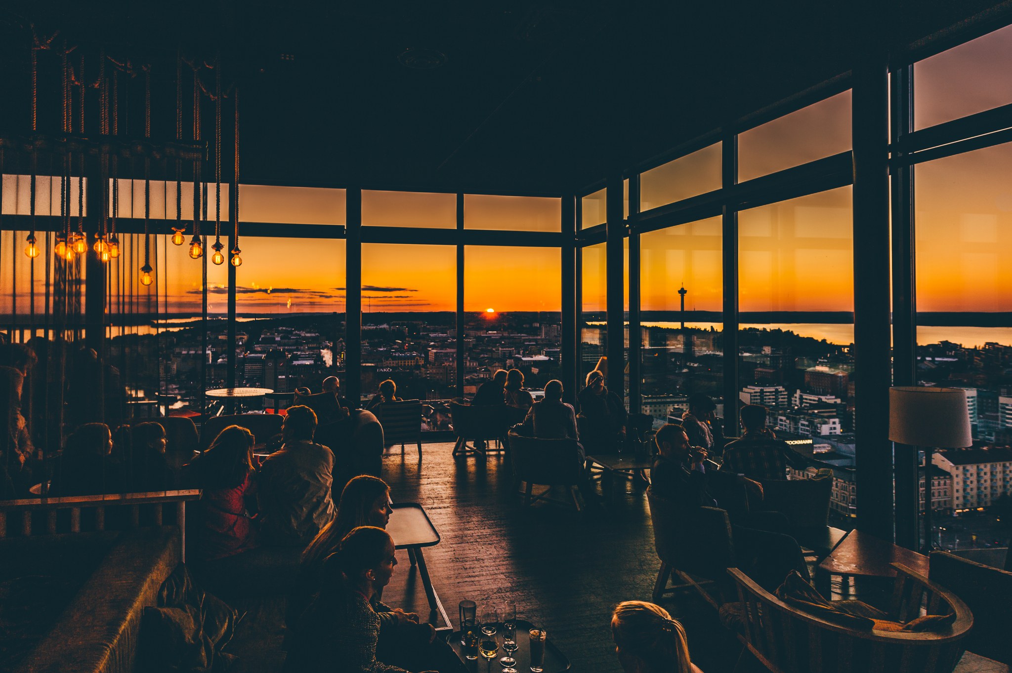 People sit inside a high-rise bar or restaurant with large windows, watching the sunset over a city landscape and distant water.