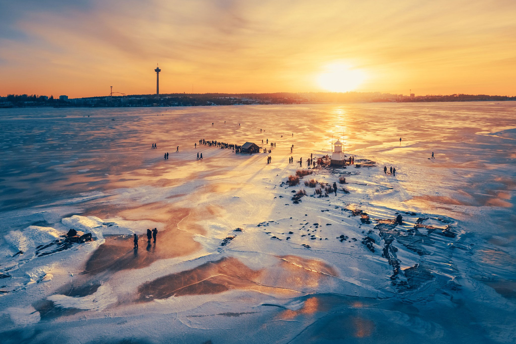 Aerial view of people walking on a frozen Lake Näsijärvi at sunset near Siilinkari Lighthouse, with the distant city skyline of Tampere in the background.