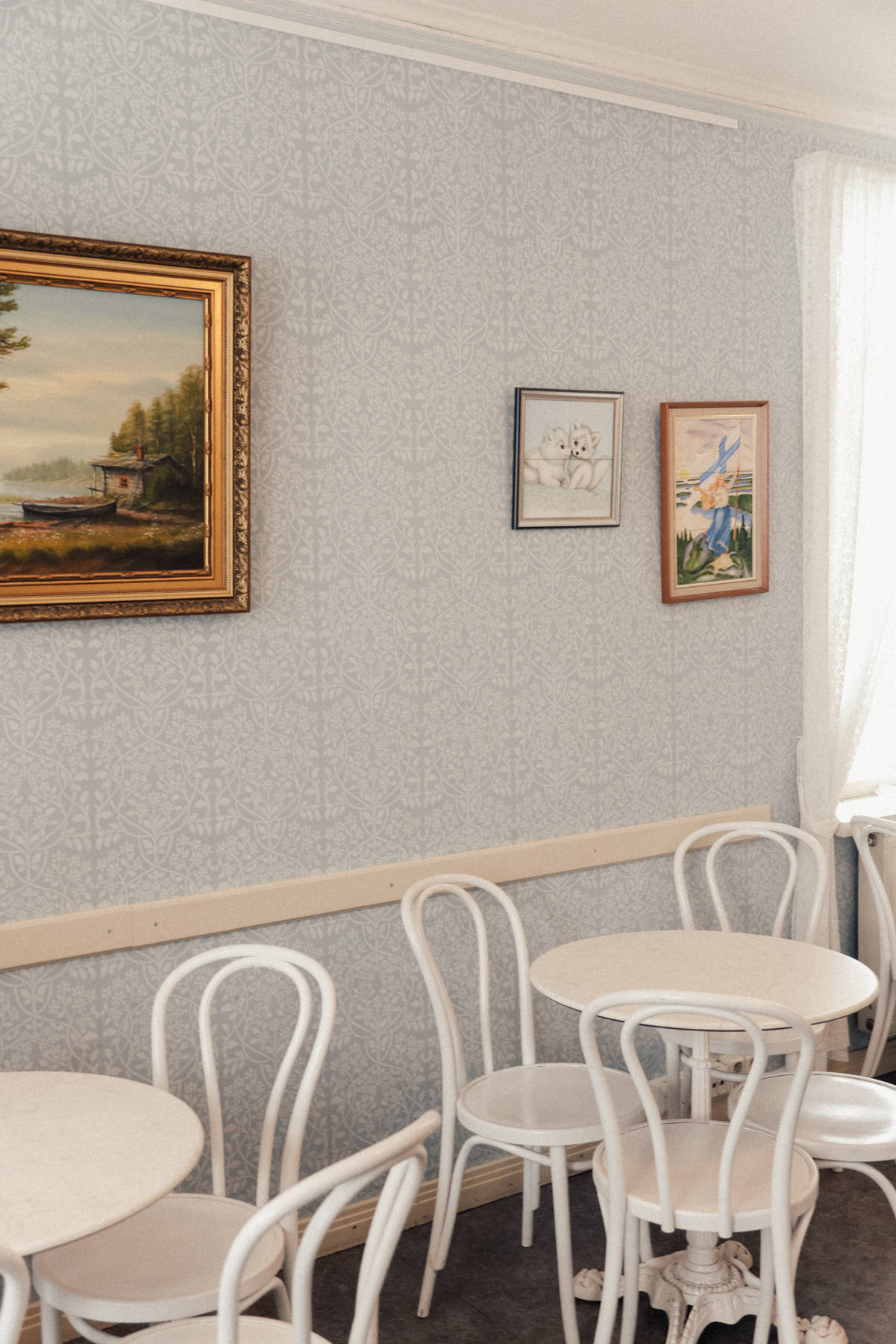 A small dining area with white round tables and chairs against a light blue wallpapered wall greets visitors Inside the Tallipihan kahvila cafe, known for its traditional Finnish food. Three framed pictures, including landscape and floral paintings, hang on the wall.