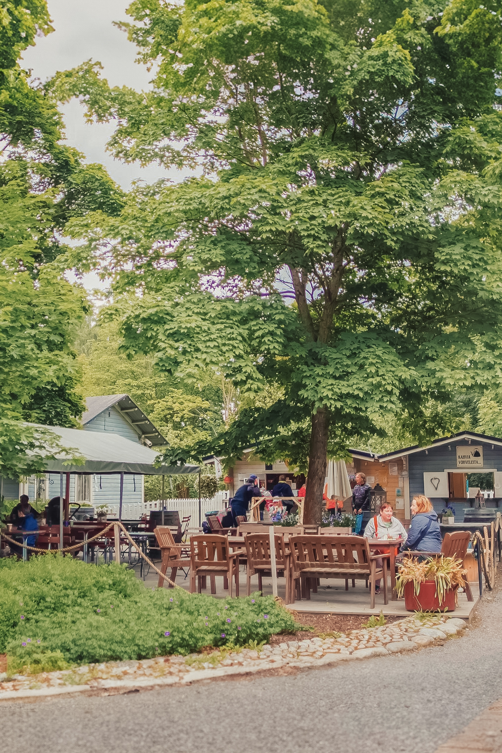 Outdoor cafe with multiple people sitting at tables under large trees near small buildings. It’s a sunny day, and the scene is lush with greenery, reminiscent of the charm found at Tallipiha Stable Yards.