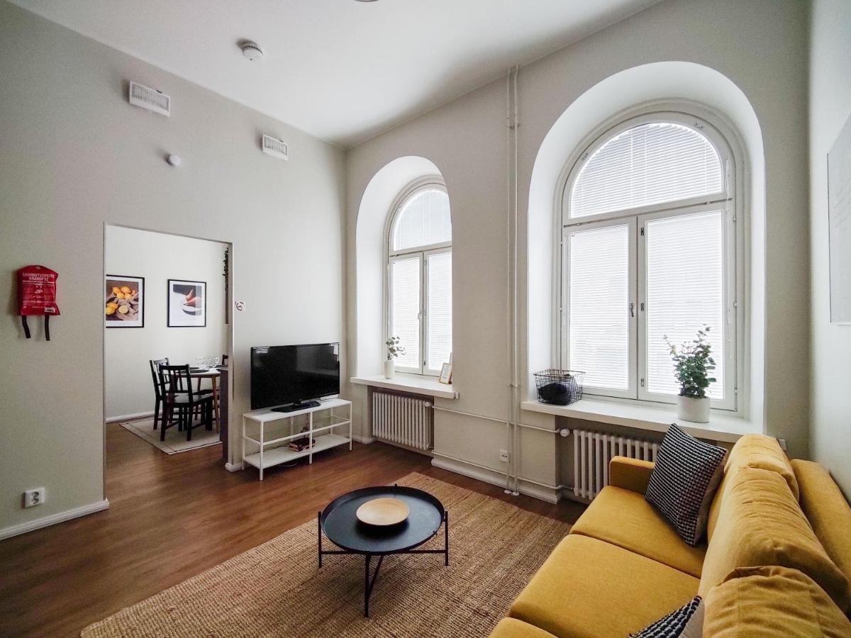 A bright living room with a yellow sofa, a TV on a stand, two large arched windows, and a round coffee table. A dining area is visible through an open doorway. Ideal for guests of the best hotels in Tampere, this cozy space offers all the comfort and charm of home.