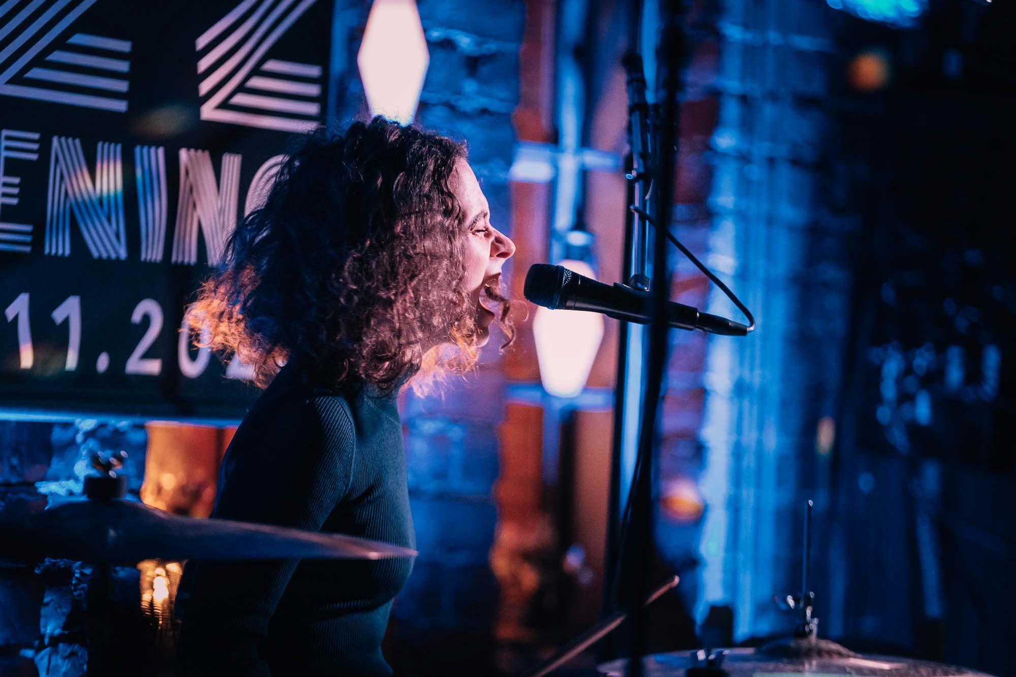 Amanda Blomqvist, with her curly hair, sings into a microphone while performing on stage in a dimly lit setting at the Tampere Jazz Happening 2020, one of the best festivals in Tampere.
