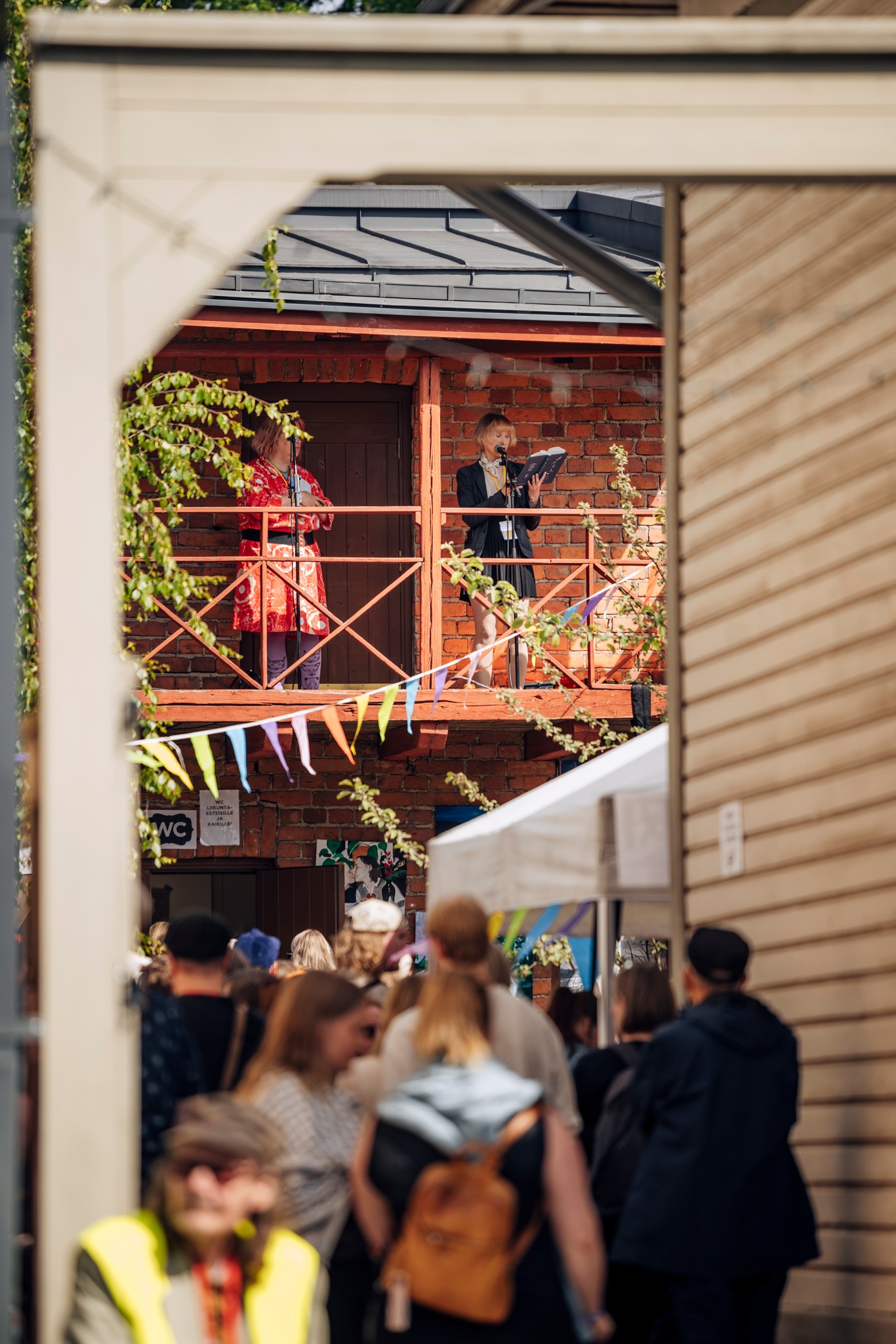 A person speaks from a balcony to a crowd below at an outdoor event, with colorful bunting and various structures in view, evoking the spirited atmosphere of the Tampera Annikki Poetry Festival.