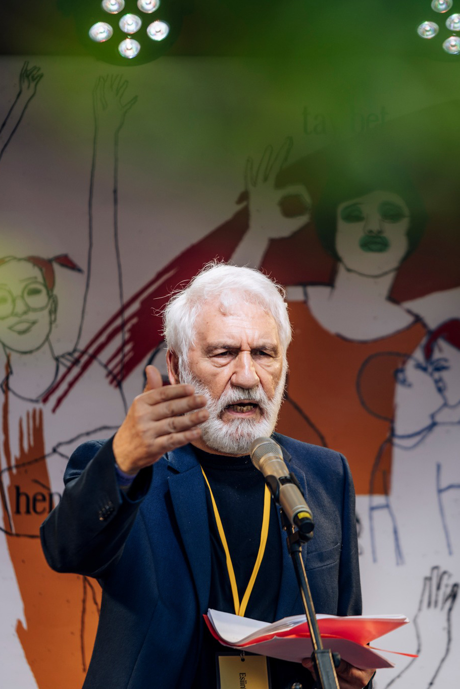 A man with a white beard speaks into a microphone, holding an open book. He gestures with his hand. In the background, there is colorful artwork depicting various figures. This scene unfolds at Annikki Poetry Festival in Tampere, where Abdulla Pashew, the most important Kurdish poet, takes center stage.