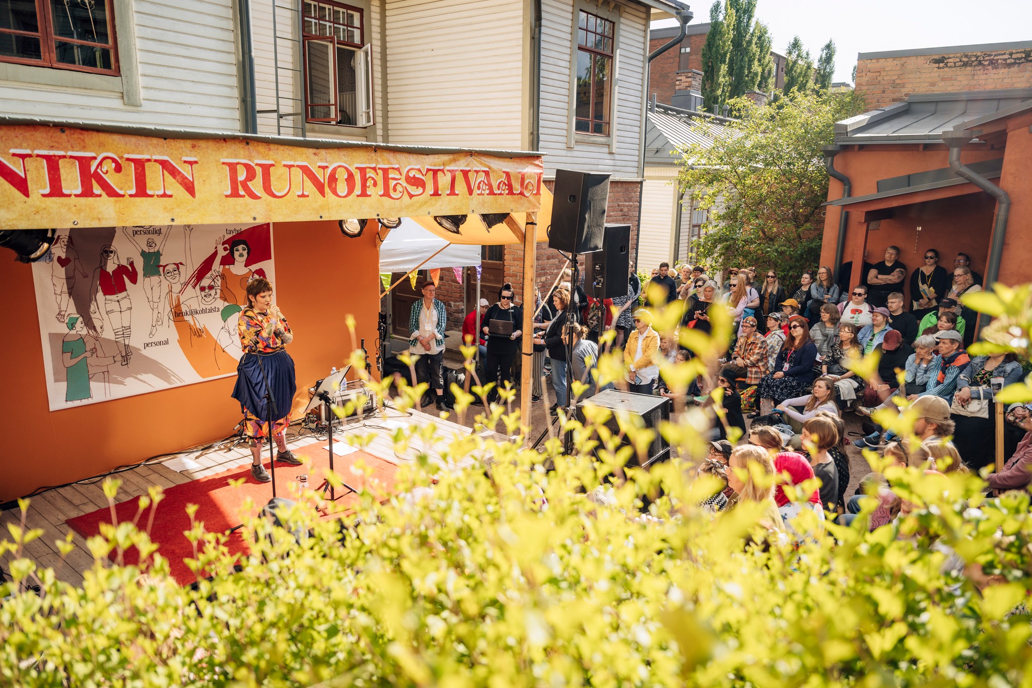 A person stands on an outdoor stage at Tampere's "Annikki Poetry Festival," passionately giving a speech with a banner reading "NIKIN RUNOFESTIVAL" overhead. An attentive audience, both seated and standing, listens intently.