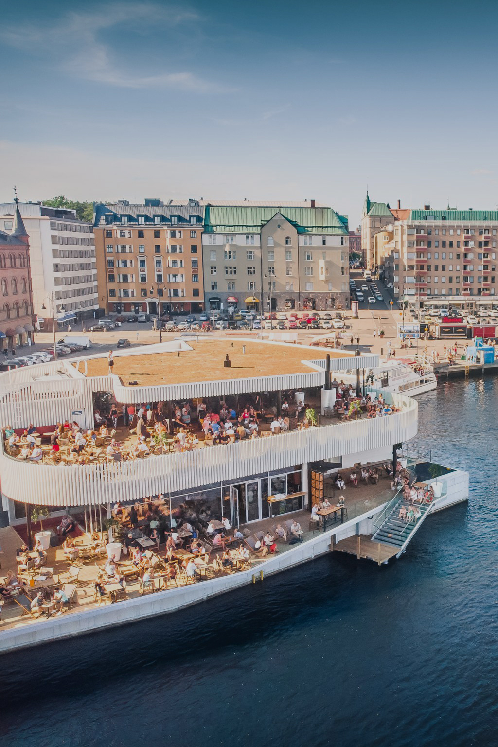 A large waterfront building with rooftop and lower deck outdoor seating is crowded with people. The building overlooks a body of water, with city buildings in the background under a clear sky.