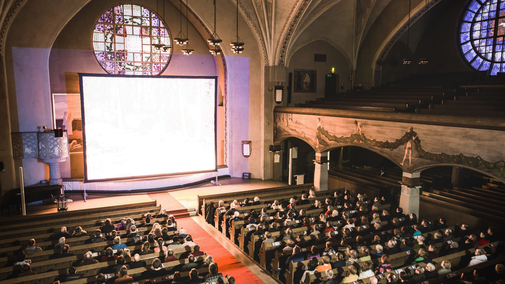 A large audience sits in a dimly lit, church-like venue at the Tampere Film Festival, with a giant projection screen at the front displaying a bright image. Stained glass windows are visible on both sides, casting colorful patterns across the room.
